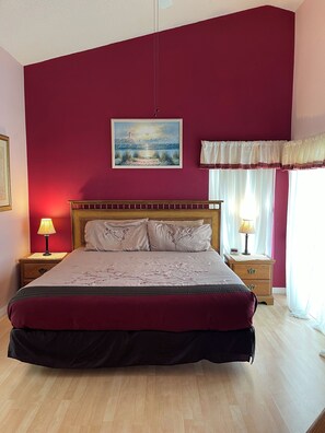 A bedroom with a large bed, two bedside tables with lamps, a painting on the wall, and a window with curtains. - Lovely 3 Bedroom 2 Bath Villa (Kissimmee)