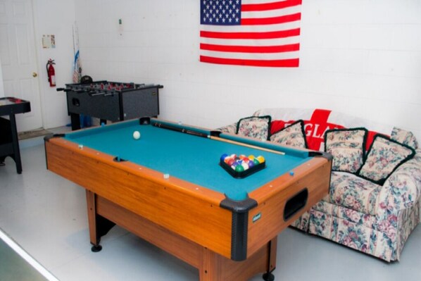 Une table de billard, un canapé à motifs floraux et un drapeau américain dans une pièce aux murs blancs.
