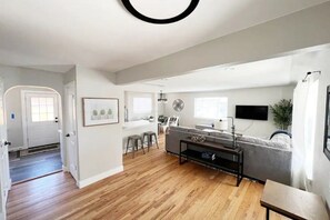 A modern living room with a grey sofa, a coffee table, and a TV. There's a dining area with a table and chairs, and a kitchen in the background. - 3 Bedroom, Open Layout - Walk to Anschutz Medical! (Aurora)