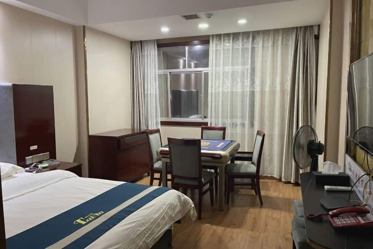 Ein Hotelzimmer mit einem Bett, einem Schreibtisch mit Stühlen, einem Fenster mit Vorhängen, einem Ventilator und einem Telefon.
