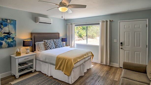 Un dormitorio con cama, mesita de noche, lámpara, equipo de aire acondicionado, ventilador de techo, ventana con cortinas y una puerta.