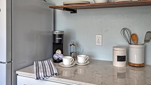 Una cocina con encimera de mármol, una cafetera Keurig, una prensa francesa, tazas blancas, una servilleta a rayas, un frasco de especias y una cuchara de madera en un recipiente blanco.