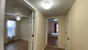 Ein Flur mit Holzboden, weißen Wänden und einer Deckenleuchte. Eine Tür führt in ein Schlafzimmer mit einem Bett und einem Fenster mit Vorhang.