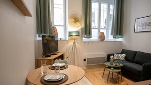 A modern living room with a dining area, featuring a sofa, a TV, a lamp, and a framed cityscape. - Opéra 2 - Lovely studio Hôtel de Ville (Lyon)