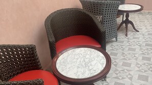 Un área de estar con sillas de mimbre y mesas redondas, con un piso de baldosas geométricas y una pared lisa.