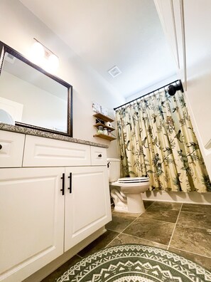 Une salle de bain avec des toilettes, un lavabo avec un miroir, un rideau de douche orné de motifs d’oiseaux et un tapis à motifs.