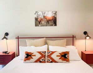 Un lit bien fait, agrémenté d’oreillers à motifs, d’une tête de lit et d’un tableau représentant des chevaux accroché au mur.