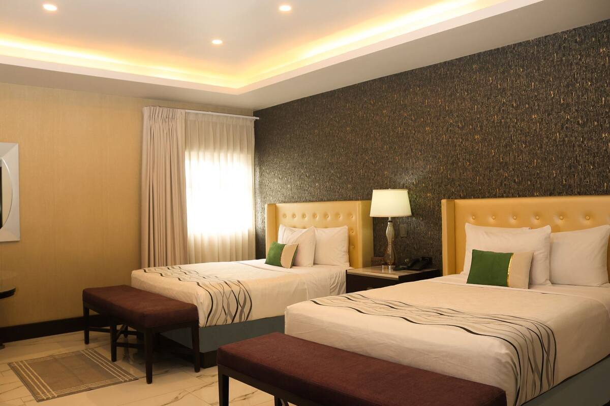 A hotel room with two beds, a nightstand with a lamp, a bench, and a large window with curtains.