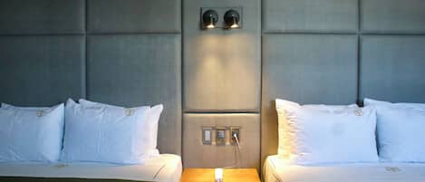 Habitación de hotel con dos camas, cabecero acolchado, mesita de noche con teléfono e iluminación en la pared.