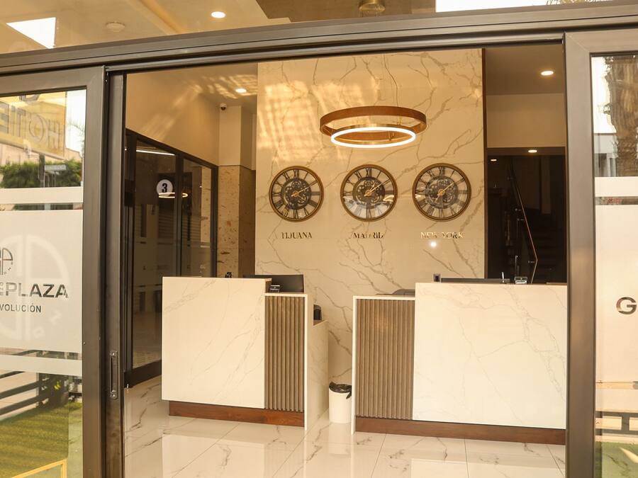 Zona de recepción con paredes de mármol, tres relojes que marcan la hora de Tijuana, Madrid y Nueva York, y un mostrador de recepción moderno.