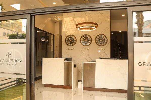 Zona de recepción con paredes de mármol, tres relojes que marcan la hora de Tijuana, Madrid y Nueva York, y un mostrador de recepción moderno.