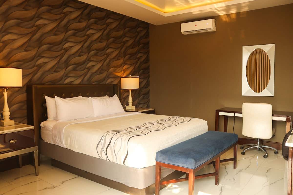 A hotel room with a bed, bedside tables, a desk with a chair, an air conditioning unit, and a decorative wall panel.
