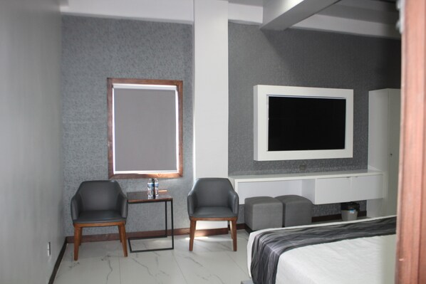 Une chambre d’hôtel moderne équipée d’un lit, de deux fauteuils, d’une petite table, d’un téléviseur fixé au mur et d’une fenêtre avec des stores.