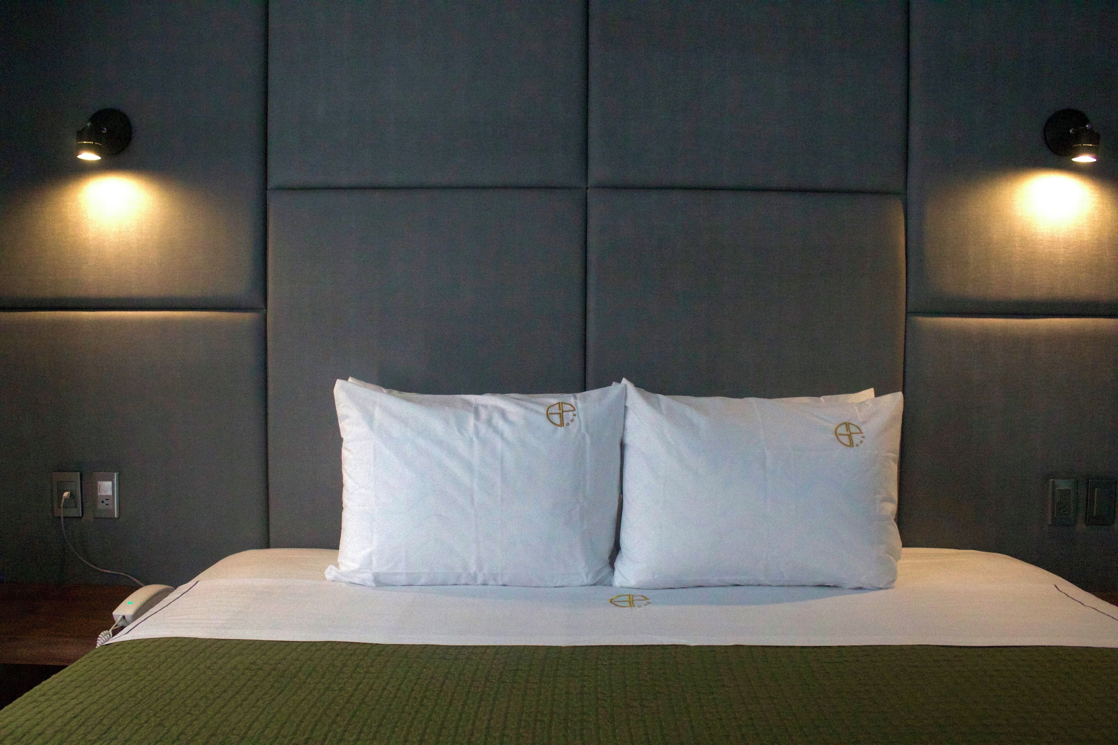 Habitación de hotel con una cama grande, dos almohadillas blancas con un logotipo, una colcha verde e iluminación instalada en la pared.