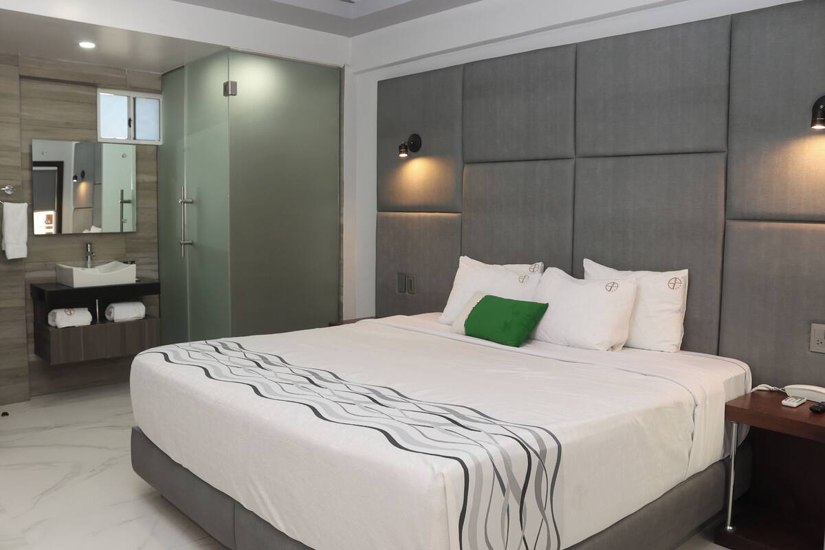 A modern hotel room with a large bed, a bedside table, a nightstand, a built-in vanity with a mirror, and a shower area.