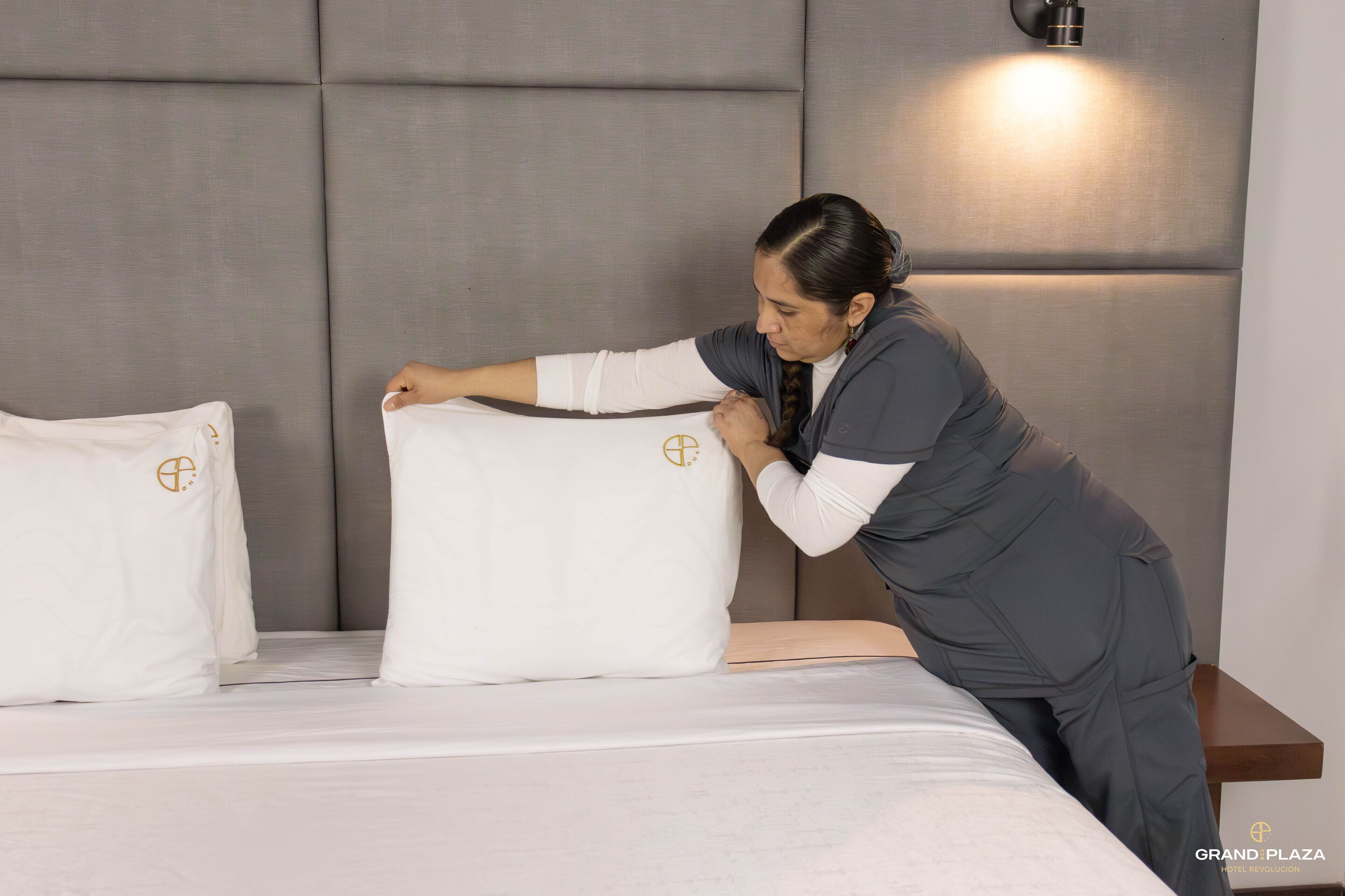 Uma pessoa ajustando um travesseiro grande e branco em uma cama com cabeceira acolchoada e luminária fixada na parede.