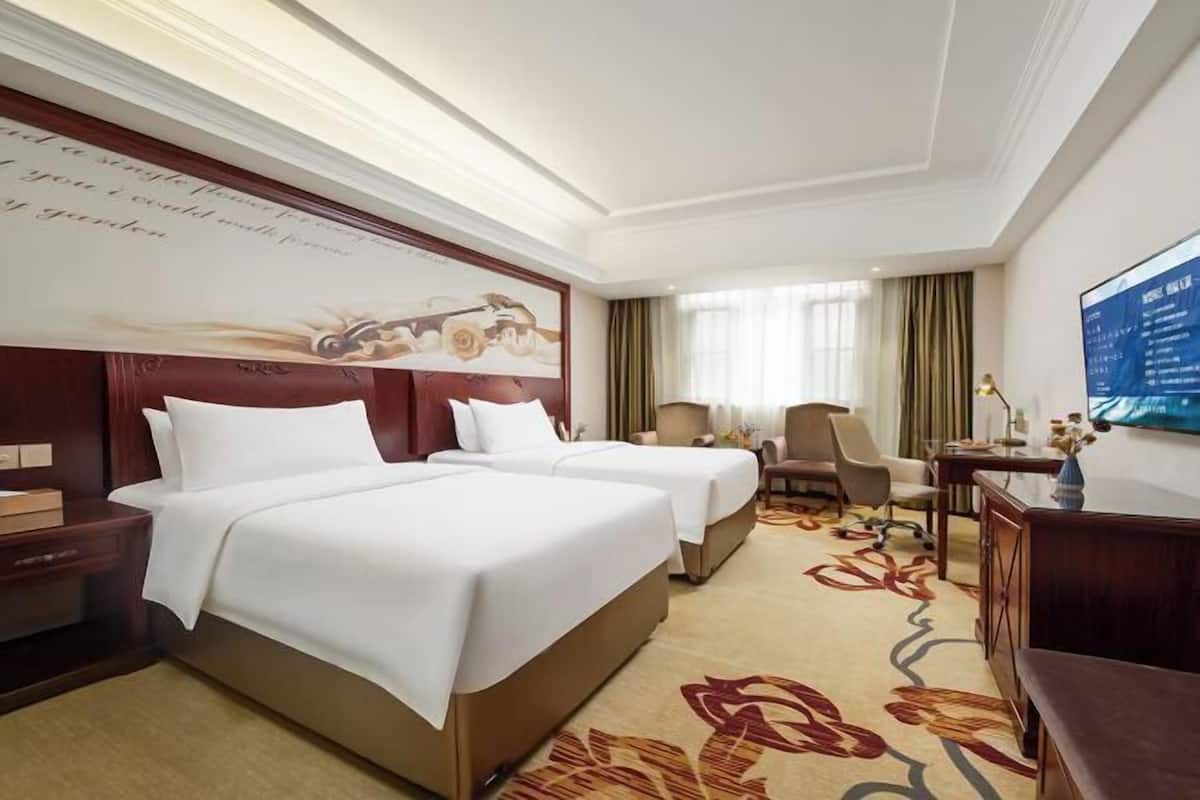 A hotel room with two beds, a desk, a chair, and a large wall-mounted TV.