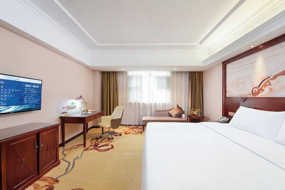 A hotel room with a bed, a desk with a lamp, a chair, a television, and a large window with curtains.