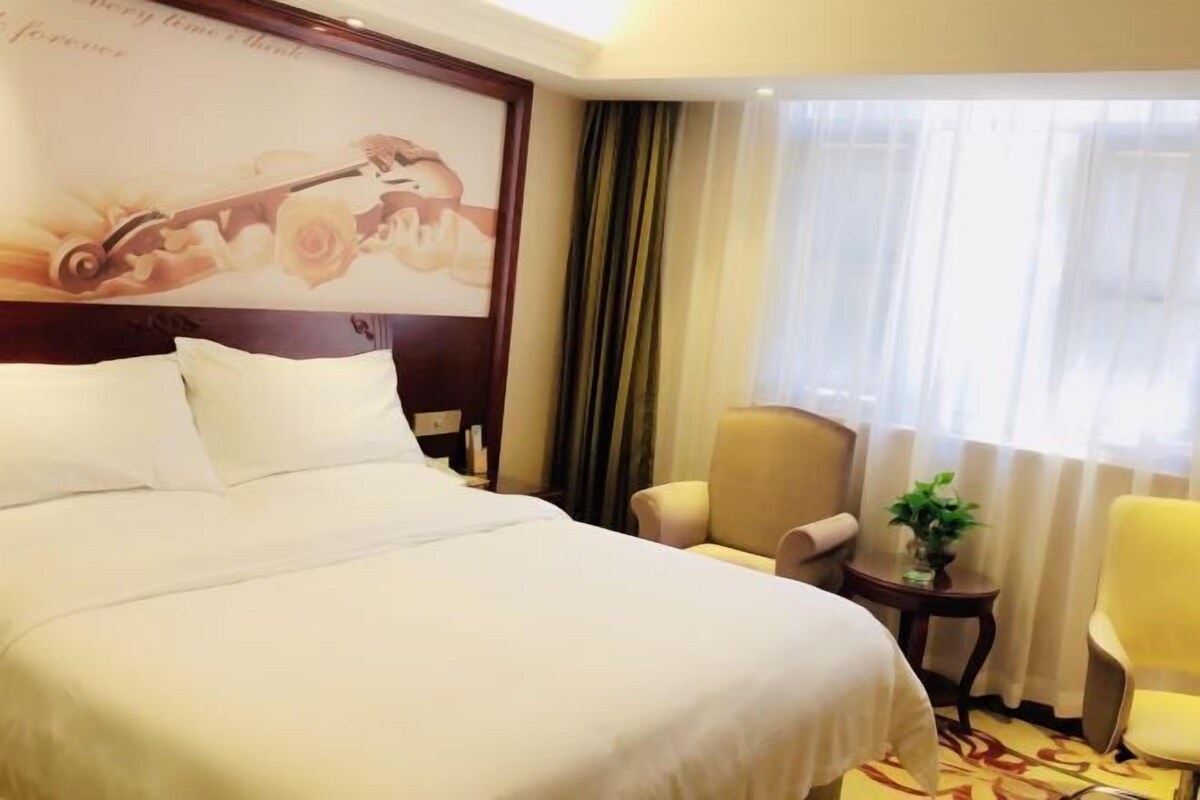 A hotel room with a large bed, a chair, a small table with a plant, and a window with curtains.