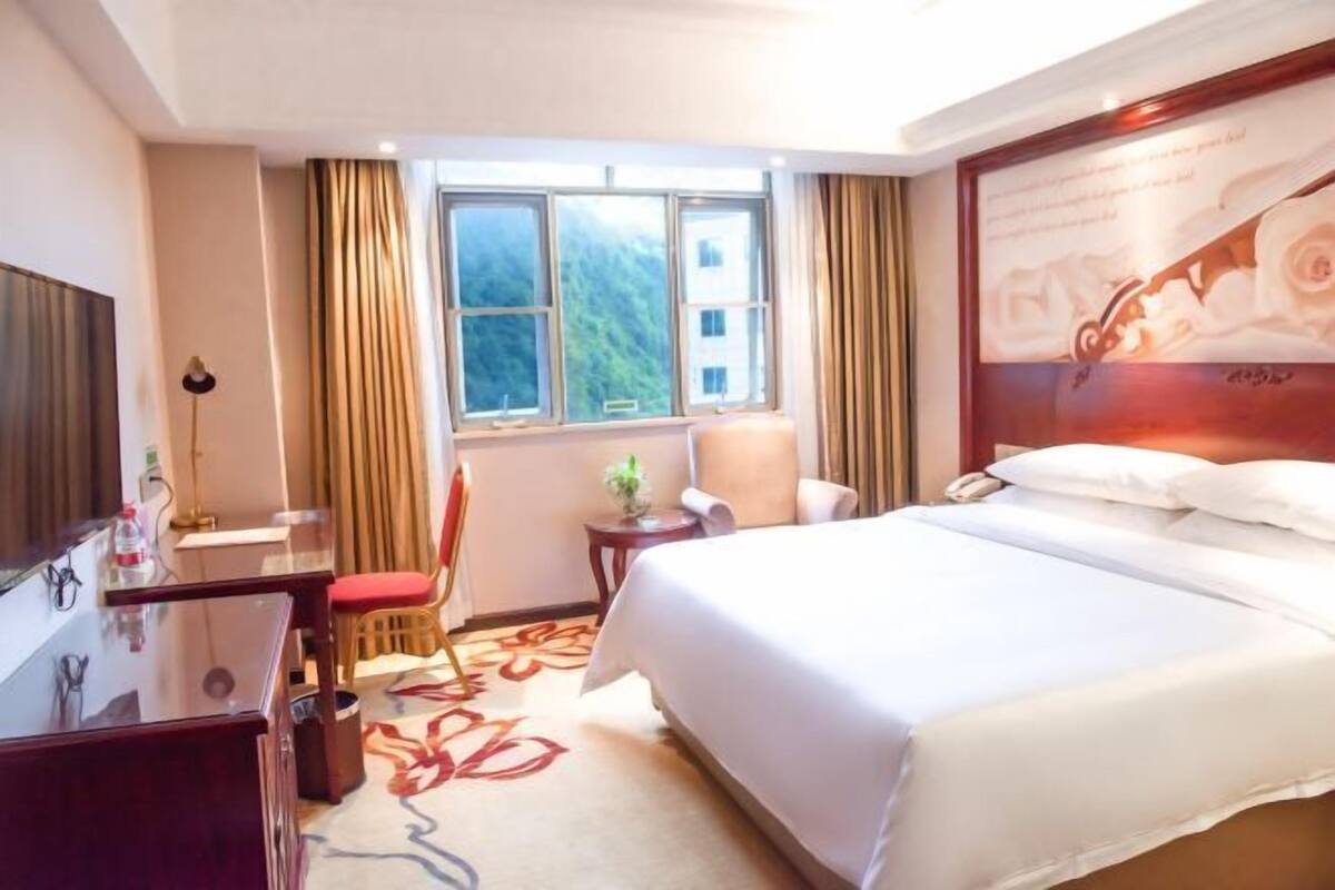 A hotel room with a bed, a desk, a chair, a window with curtains, and a wall-mounted TV.