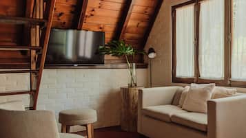 A cozy room with a wooden ceiling, a flat-screen TV mounted on the wall, a white brick wall, a beige sofa, a small wooden stool, and a potted plant.