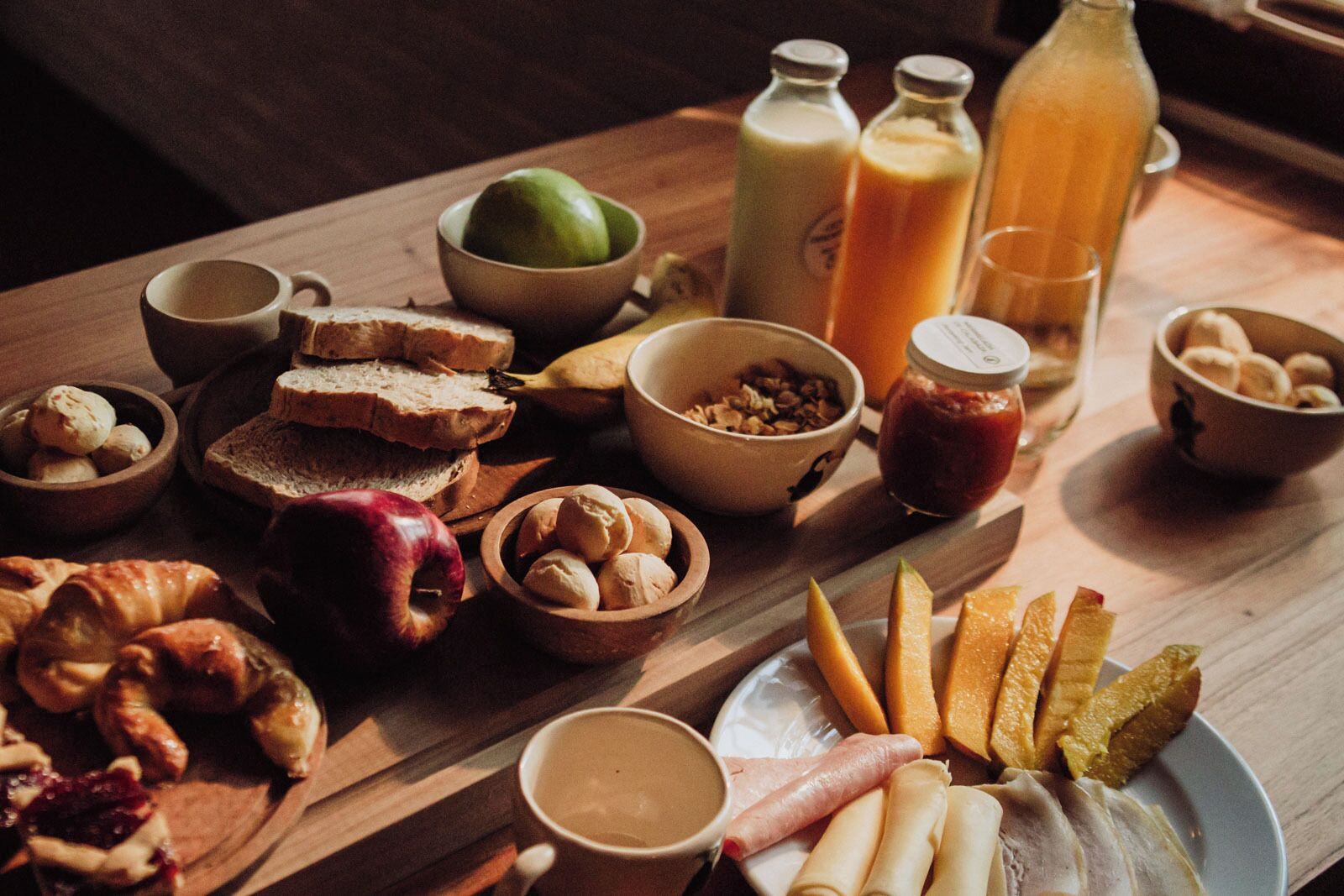 Una mesa de madera con diversos productos para el desayuno, como pan, bollería, frutas y bebidas.