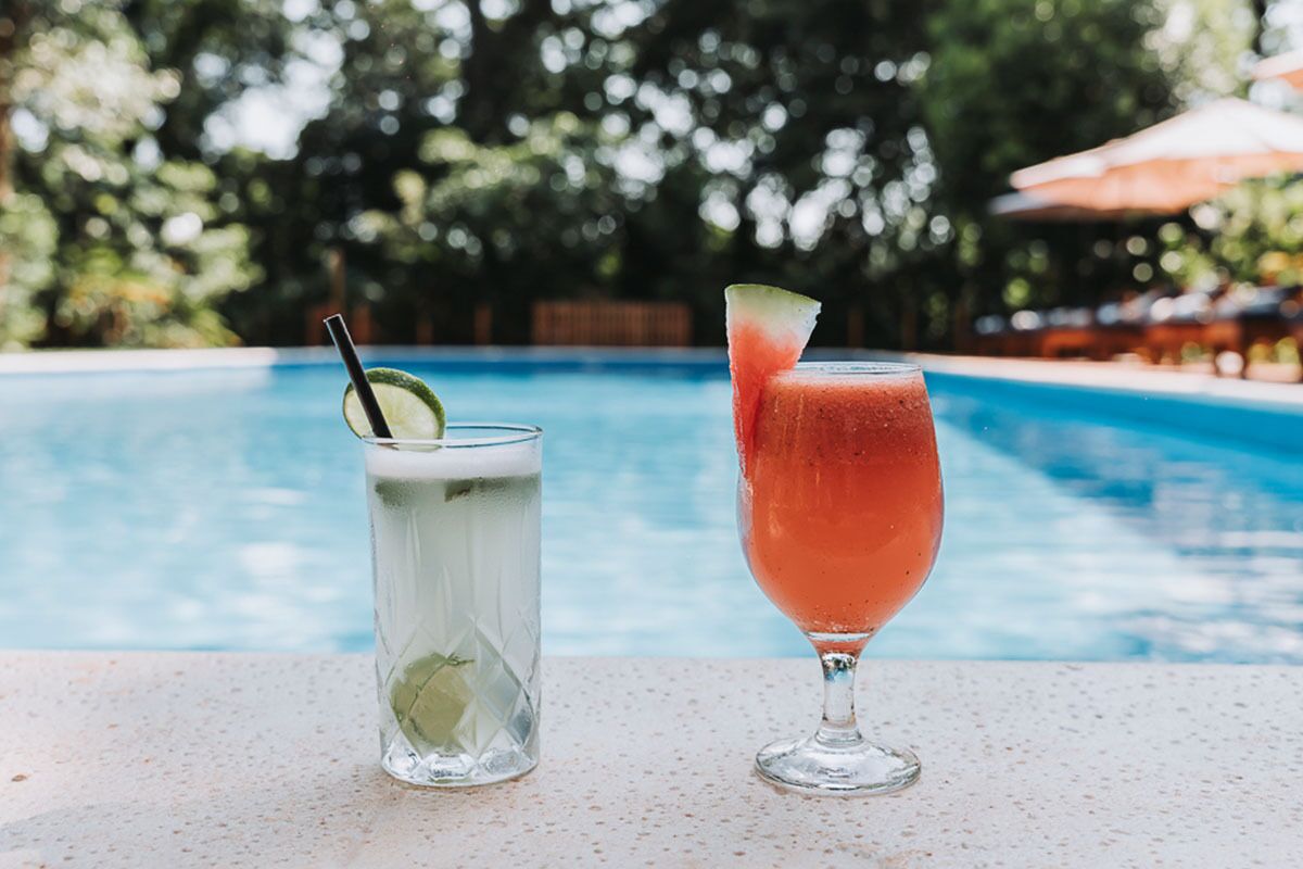 Deux boissons posées au bord d’une piscine, avec un arrière-plan flou d’arbres et de parasols.
