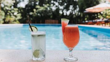 Deux boissons posées au bord d’une piscine, avec un arrière-plan flou d’arbres et de parasols.