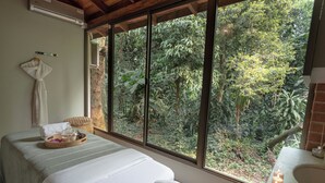 Ein Spa-Raum mit Massageliege, einem Fenster mit Blick auf den Wald und einer Badewanne mit Kerzen.