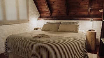 A bedroom with a wooden ceiling, a white brick wall, a striped bedspread, and a bedside table with a lamp.