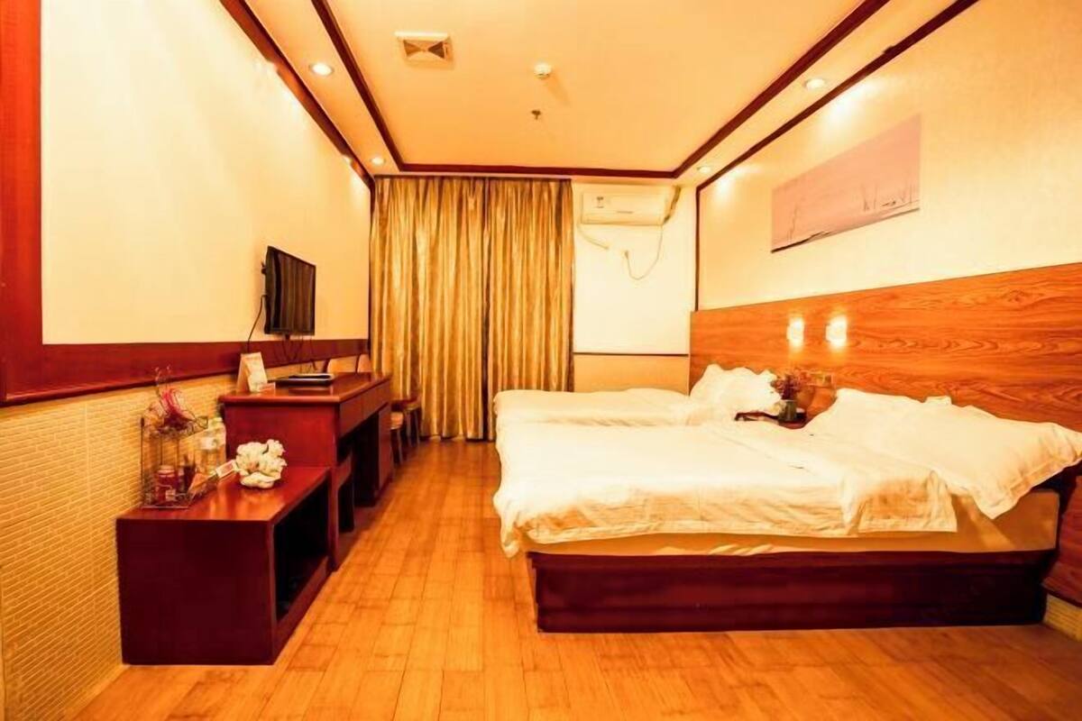 Habitación de hotel con piso de madera, cama con ropa blanca, escritorio con silla, televisión y ventana con cortinas.