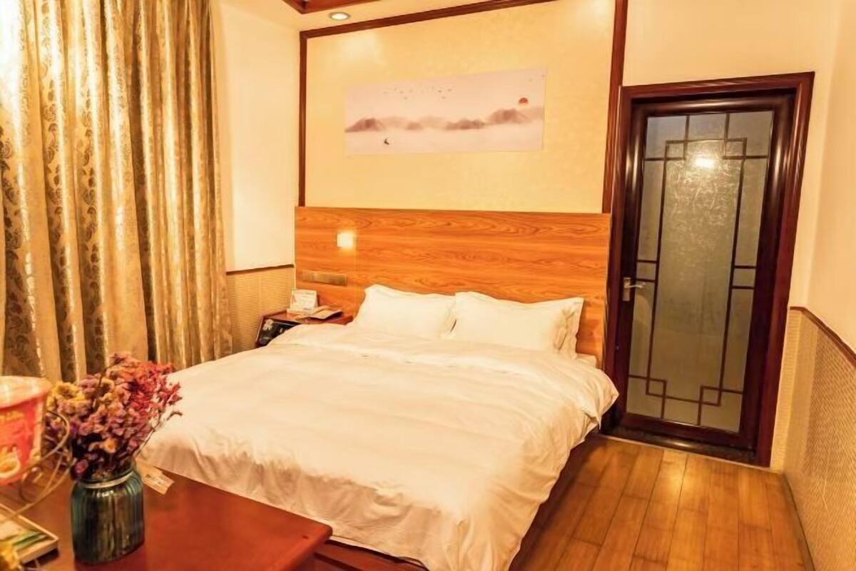 Habitación de hotel con cabecera de madera, cama con ropa blanca, mesita de noche con lámpara, florero con flores y puerta con panel de vidrio.
