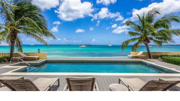 A poolside view with a clear blue sky, two palm trees, and a view of the ocean. - Encanto- 2 Bedroom Beachfront Condo - Simpson Bay! (Simpson Bay)