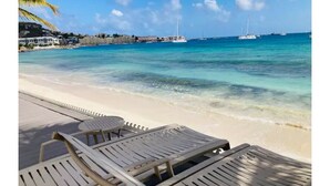 A beach with clear turquoise water, a row of lounge chairs, and a distant view of sailboats. - Encanto- 2 Bedroom Beachfront Condo - Simpson Bay! (Simpson Bay)