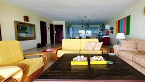 A living room with a yellow sofa, a striped sofa, a coffee table with decorative items, and a lamp. - Encanto- 2 Bedroom Beachfront Condo - Simpson Bay! (Simpson Bay)