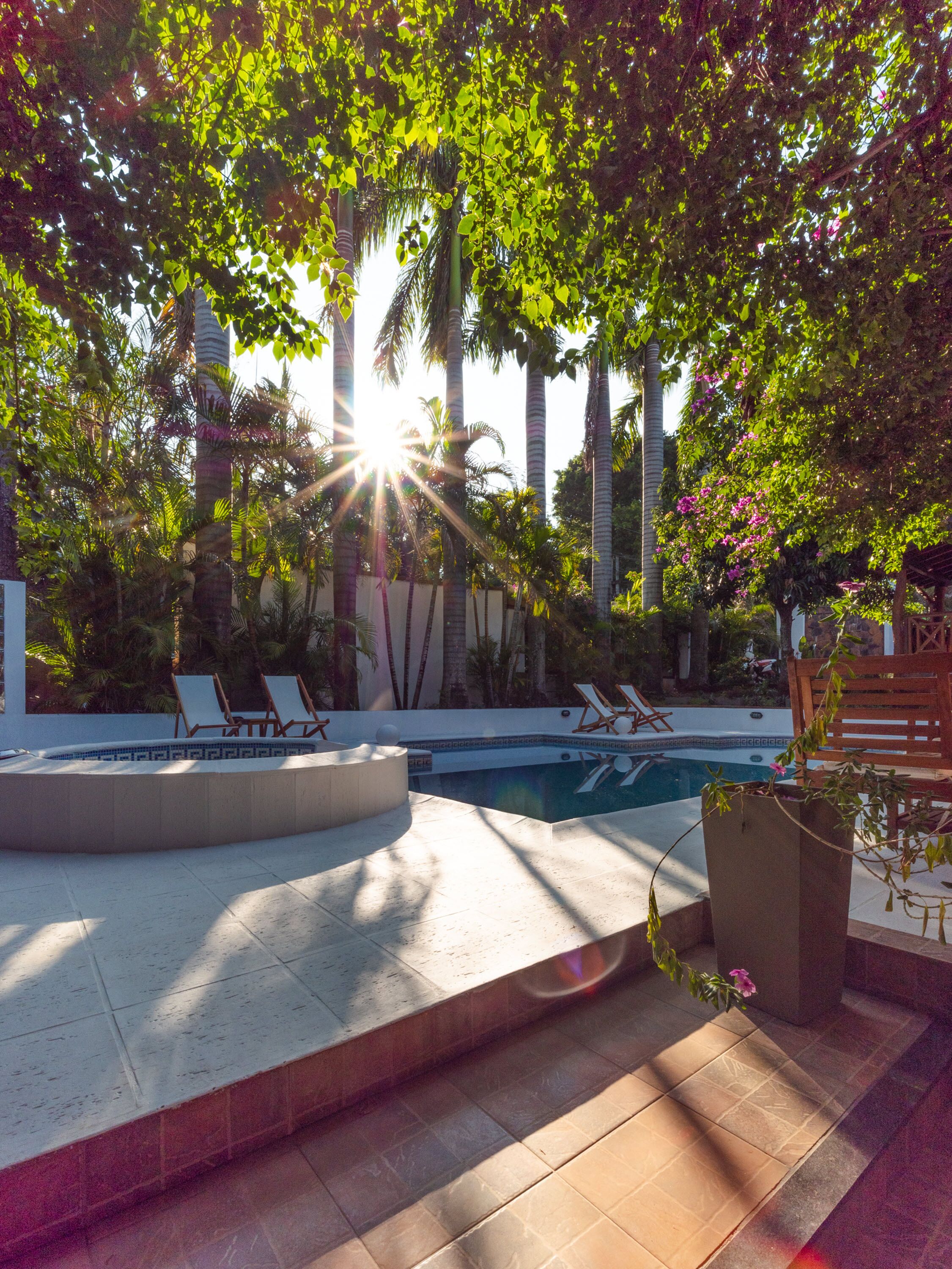 A pool area with lounge chairs and a hot tub, surrounded by palm trees and a pergola.