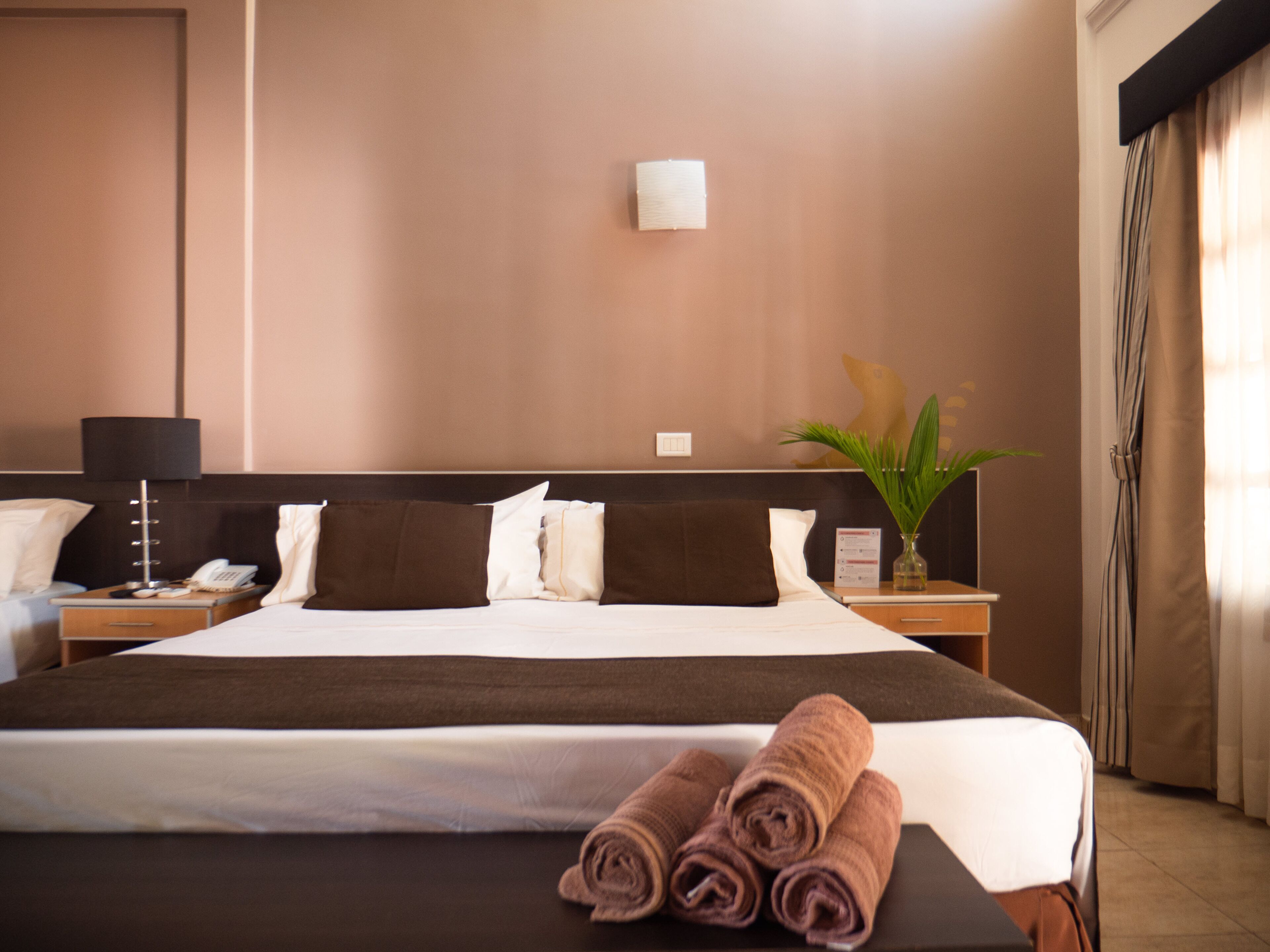A hotel room with a large bed, a nightstand with a lamp, a vase with a plant, and a window with curtains.