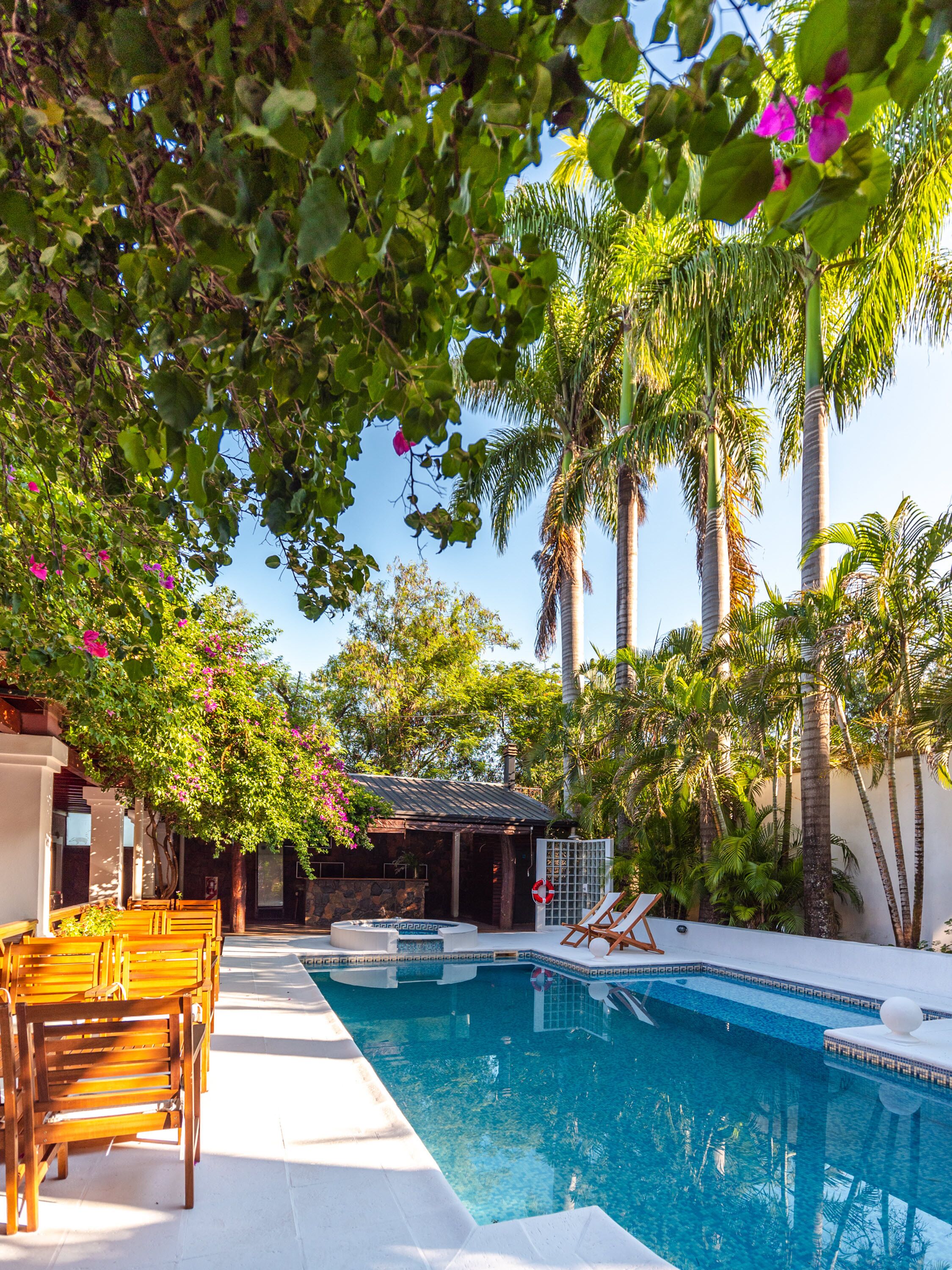 A swimming pool with a wooden deck and lounge chairs, surrounded by palm trees and flowering plants.