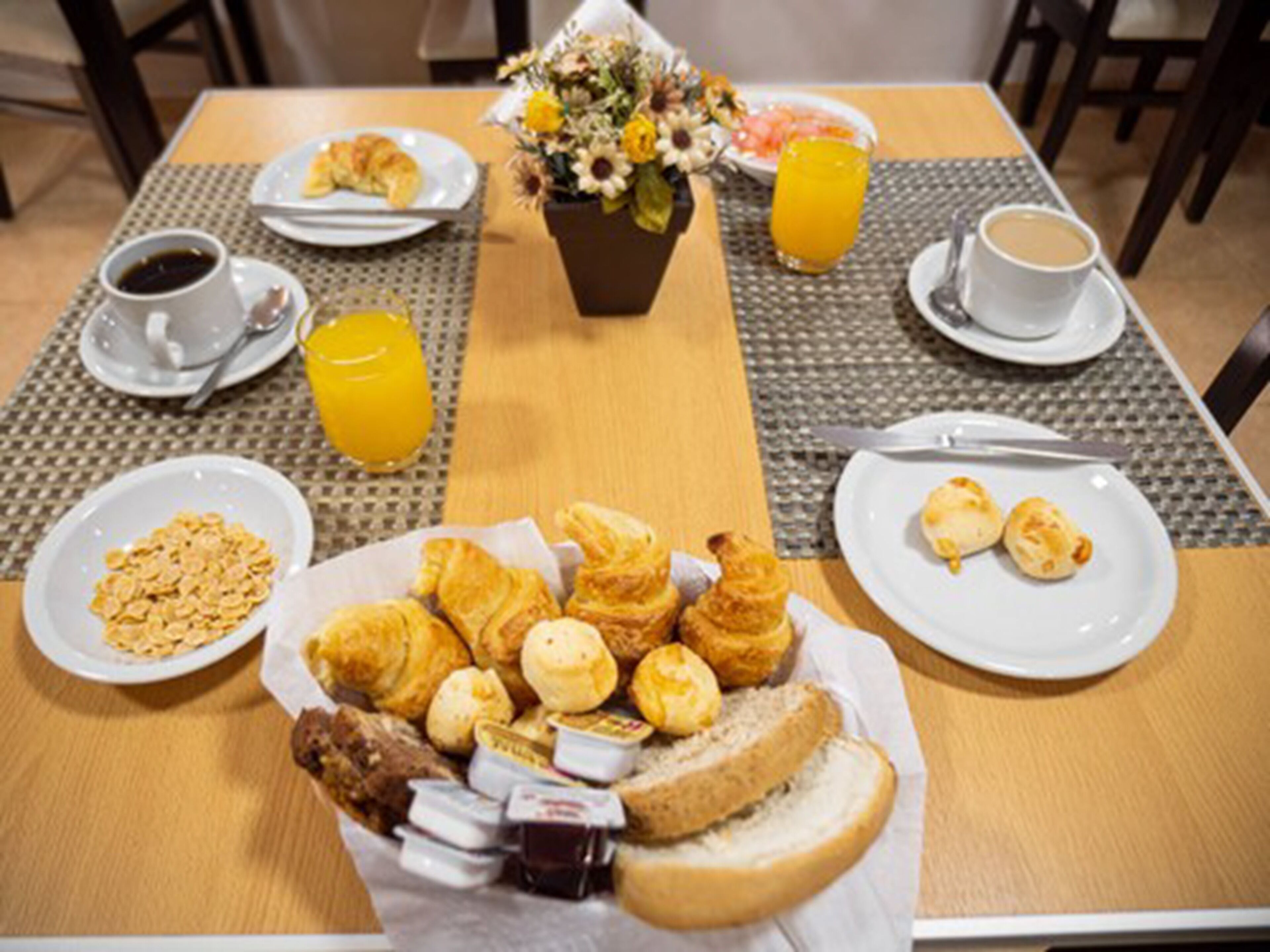 Une table de petit-déjeuner avec des croissants, des viennoiseries, du pain et un saladier de fruits. On y trouve des tasses de café et du jus d’orange, ainsi qu’un vase rempli de fleurs.