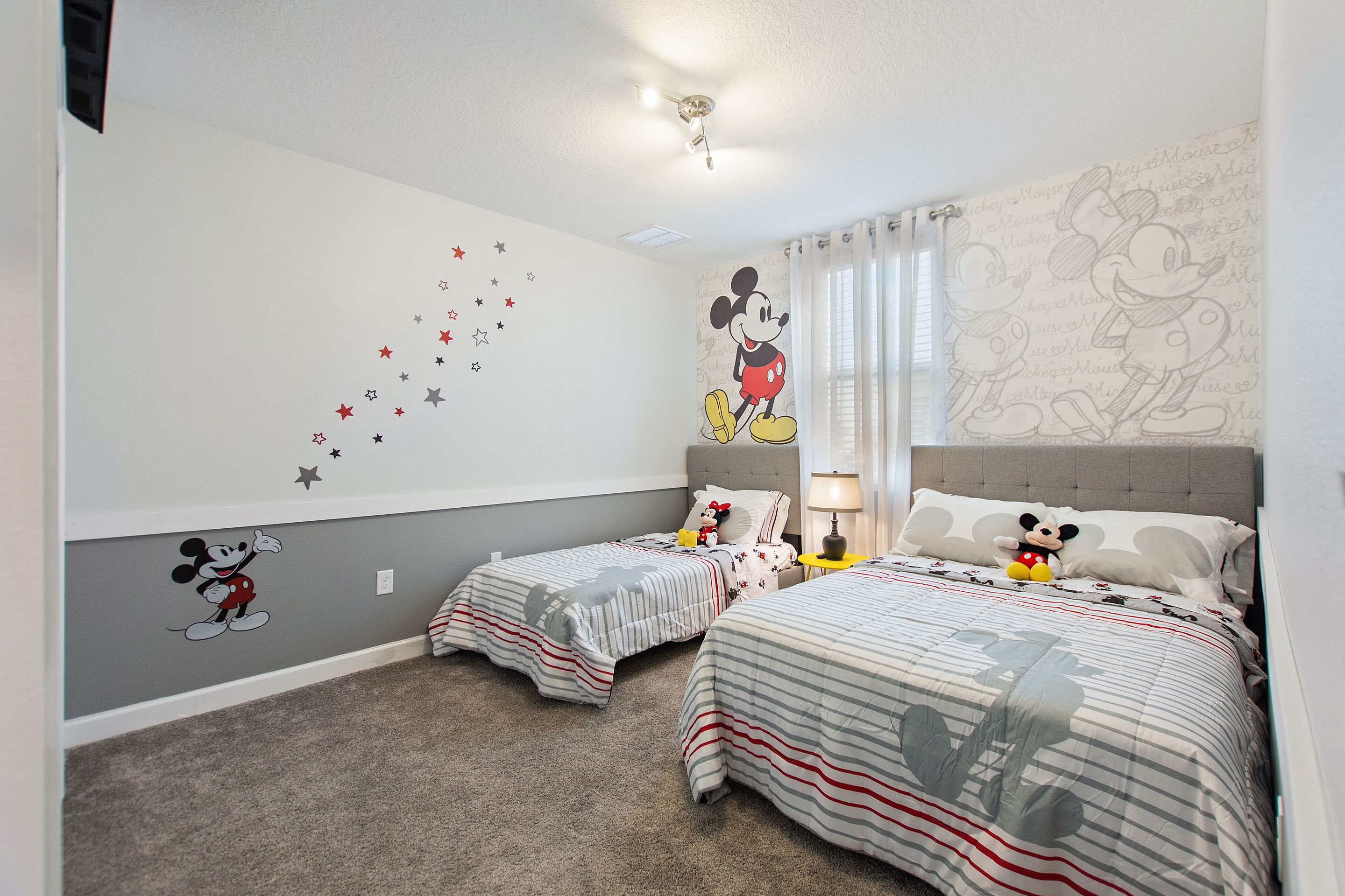 A bedroom with two beds, Mickey Mouse wall decals, and a ceiling light fixture.