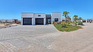 A modern building with two garage doors, a paved driveway, and landscaped areas with palm trees and flowers. - Beach Front House - EV charging included (Son.)