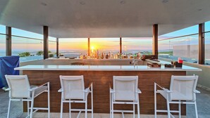 A modern bar area with a long counter, multiple bar stools, and a view of the sunset over the ocean. - Beach Front House - EV charging included (Son.)