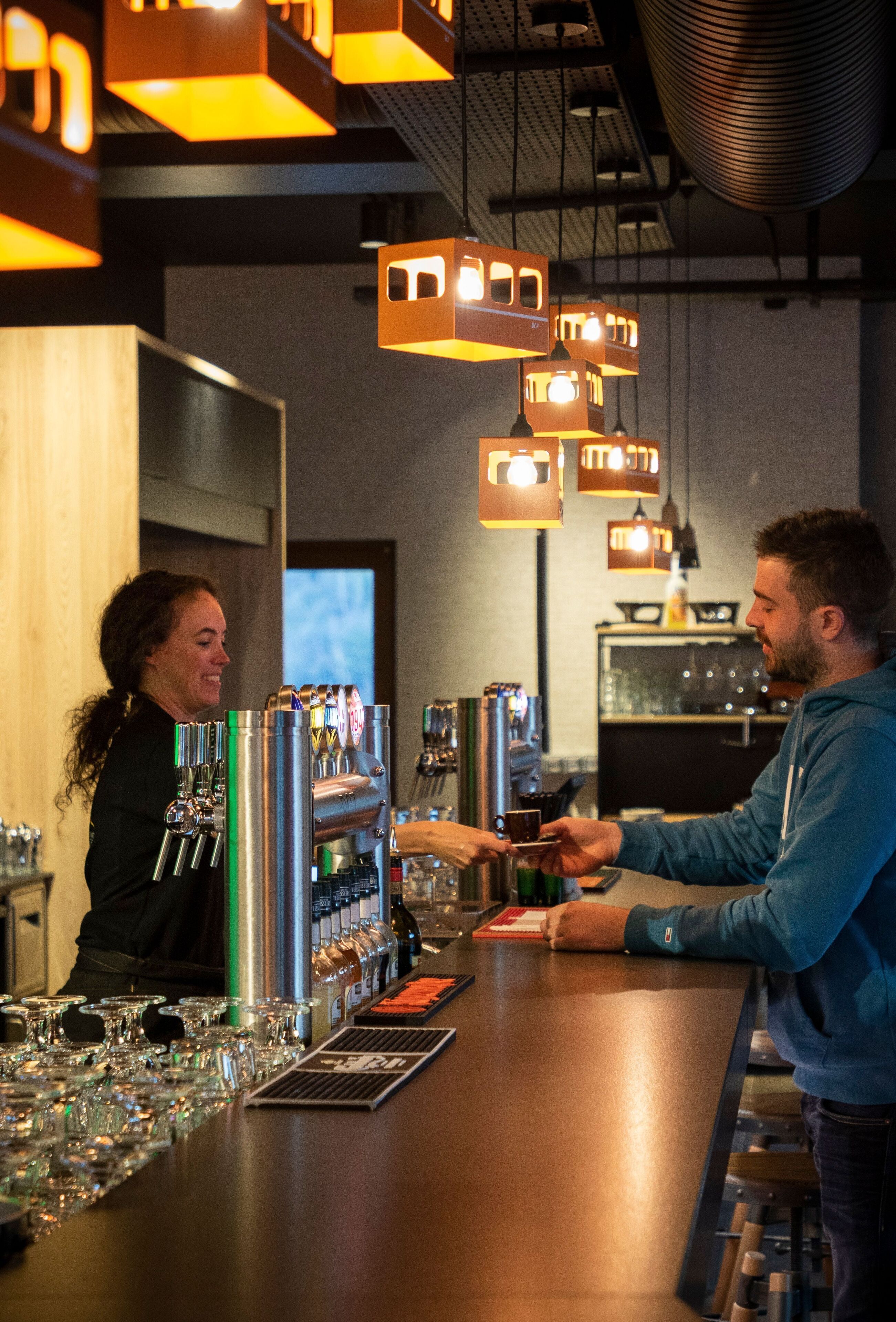 Un espace bar avec un barman et un client, des étagères remplies de bouteilles et des lampes suspendues.