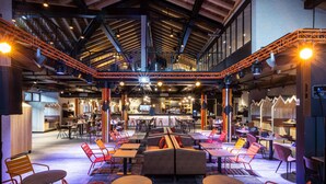 En spiseplads indendørs med flere borde og stole, en bar i baggrunden og et højt loft med synlige bjælker.