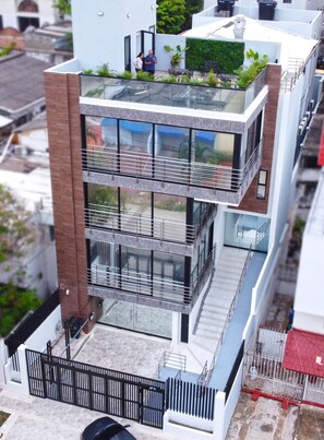 Un immeuble moderne à plusieurs étages, doté d’un balcon en verre à l’étage supérieur, d’une rambarde en métal et d’un petit espace jardin.
