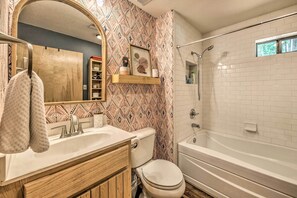 A bathroom with a wooden vanity, a white sink, a toilet, and a bathtub with a showerhead. - 'Inn the Clouds' - Charming Cloudcroft Escape (Cloudcroft)