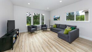 Ein geräumiges Wohnzimmer mit einer grauen Eckcouch, zwei Sesseln, einem Fernseher auf einem schwarzen Standfuß und einem Bücherregal. Große Fenster sorgen für natürliches Licht und Blick ins Grüne.