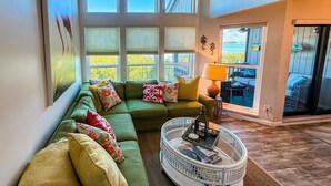 A green sectional sofa with multiple cushions, a glass coffee table with decorative items, and a view of a water body through large windows.