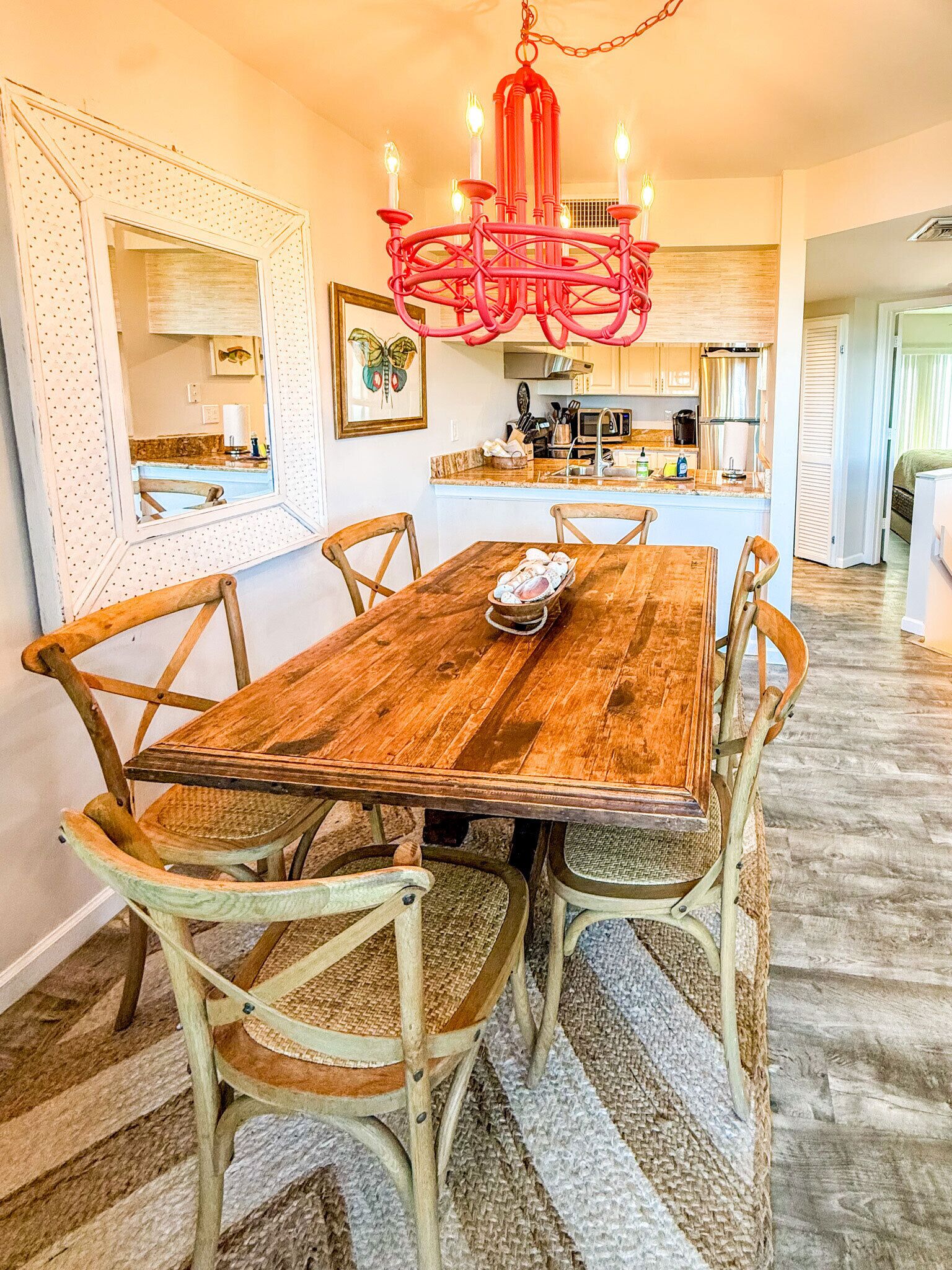 Un coin repas avec une table en bois, six chaises, un lustre rouge et, au fond, une cuisine.