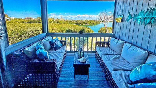 A covered porch with wicker furniture, a ceiling fan, and a view of a river and greenery.